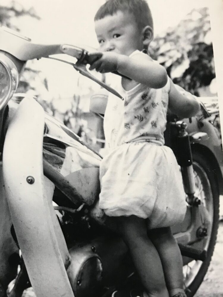 モノクロ写真。カブのようなバイクのハンドルを握り、カメラに目線を送る。幼少期の山本先生